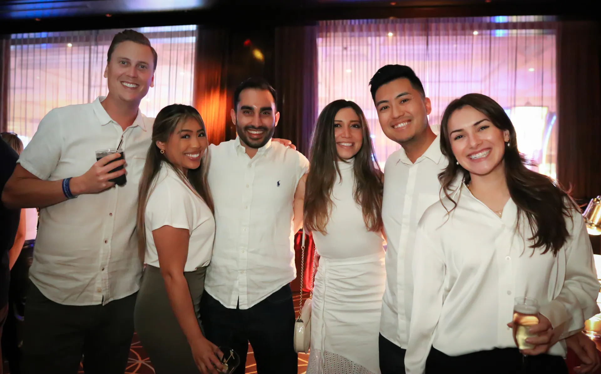 Group of six smiling young adults dressed in white shirts posing together indoors at a social event.